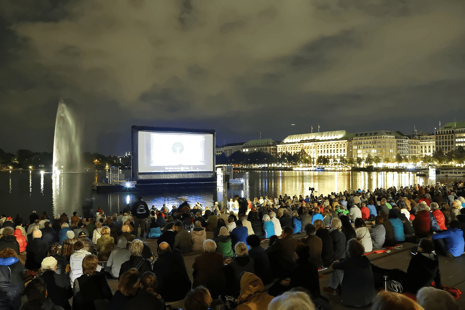 Eine Kinoleinwand auf der Alster, davor sitzen Menschen auf den Treppen am Jungfernstieg