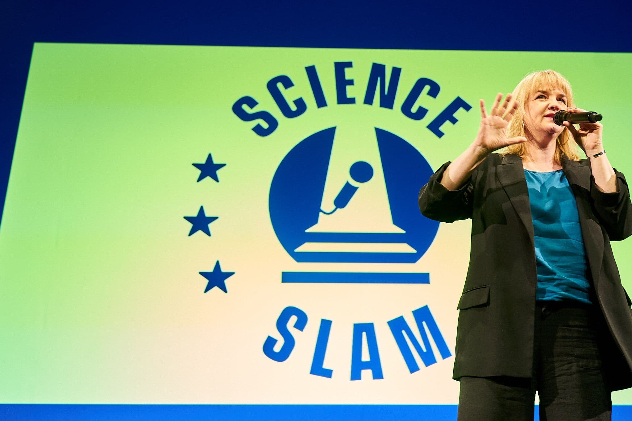 Foto einer Frau auf einer Bühne mit einem Mikrofon in der Hand, vor einem Hintergrund mit der Aufschrift "Science Slam"