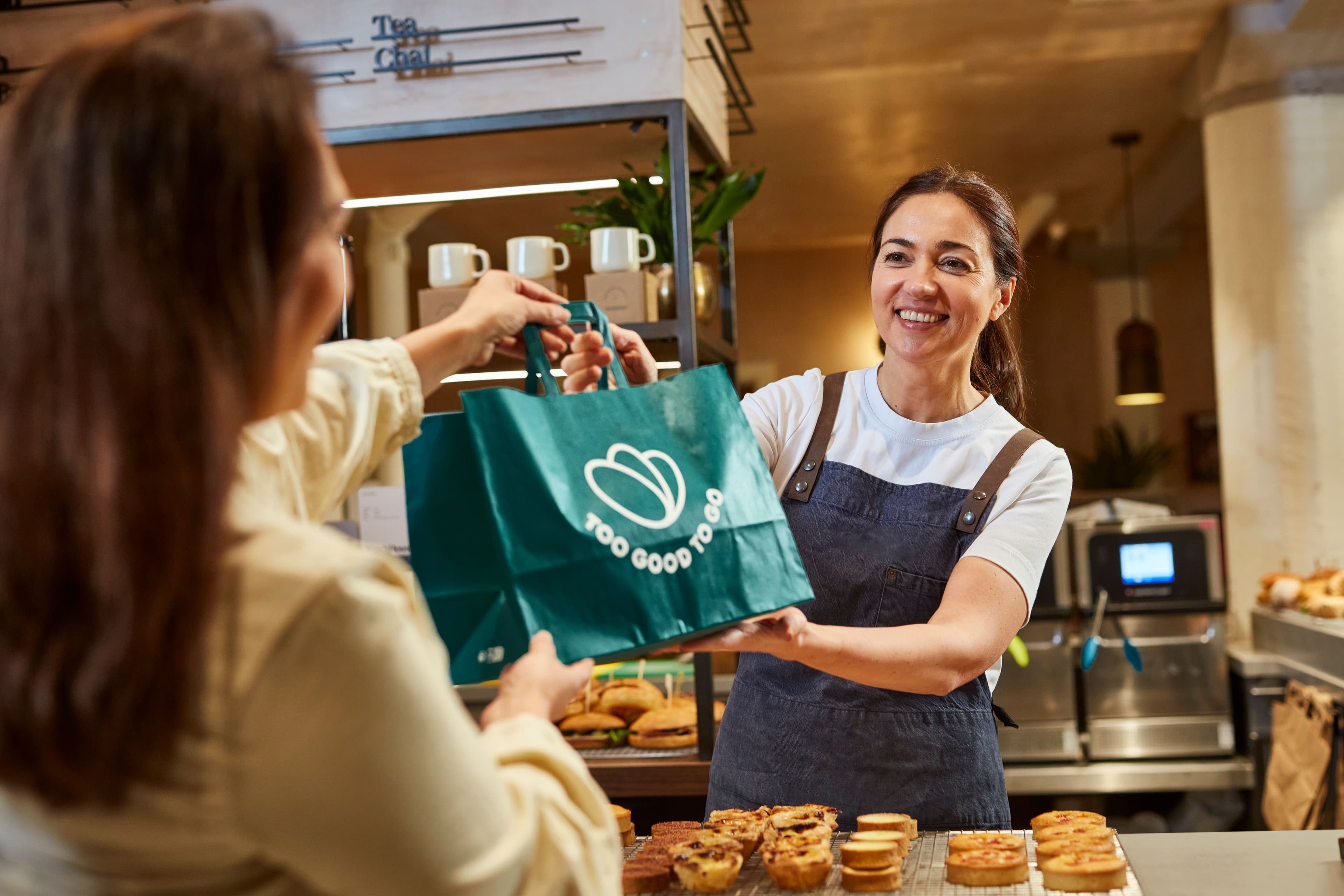 Eine Frau überreicht einer anderen Frau eine Too Good To Go Tüte in einem Geschäft